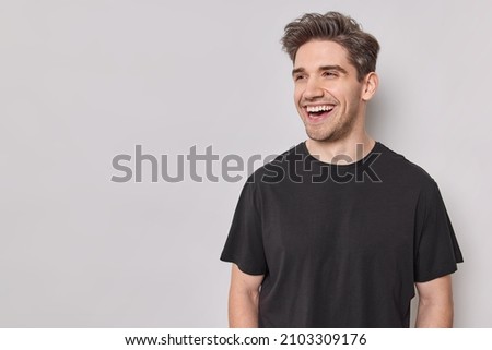 Horizontal shot of handsome cheerful man looks happily away smiles broadly wears casual black t shirt isolated over white background with blank copy space being in good mood laughs at something