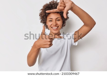 Happy woman with curly hair got inspiration imagines hot to capture interesting shot makes frame gesture smiles gladfully dressed in casual t shirt isolated over white background found great spot