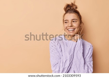 Horizontal shot of happy European woman looks dreamy away keeps hands together smiles broadly has good mood wears knitted sweater poses against beige bacground with blank copy space for promo