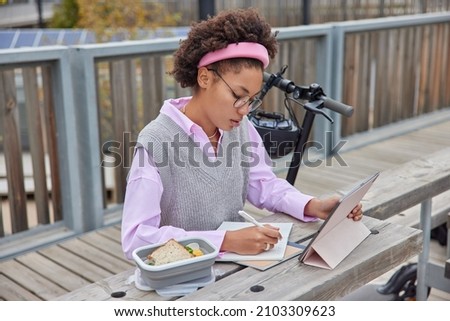 Outdoor shot of curly female student writes down impornat ideas in planner watches webinar on tablet wears neat clothes eats snack poses at wooden table near electric scooter prepares for exam