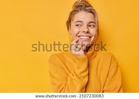 Beautiful glad woman smiles tenderly looks away with happy expression thinks about pleasant things wears casual soft jumper isolated over yellow background blank copy space for your promotion