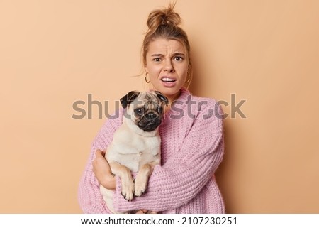 Indignant displeased young woman frowns face and looks discontent at camera hears bad news from vet about pet disease holds pedigree pug dog poses against beige background being fond of animals