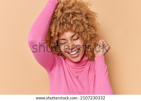Pretty joyful curly haired woman keeps eyes closed from satisfaction smiles toothily celerates something wears pink turtleneck isolated over beige backgound. Carefree female model dances indoor