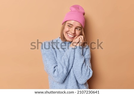 Positive dreamy European woman keeps hands near face smiles broadly being in good mood thinks about something pleasant wears hat and blue jumper isolated over brown background feels awesome.