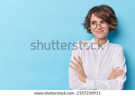 Satisfied elegant woman feels satisfied keeps arms folded considers suggestion looks away with glad expression wears round spectacles formal white shirt isolated over blue background copy space