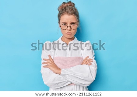 Angry woman mad as evil. Unhappy offended female model has bothered hateful expression crosses arms against chest stands in defensive pose wears spectacles and white shirt isolated on blue wall