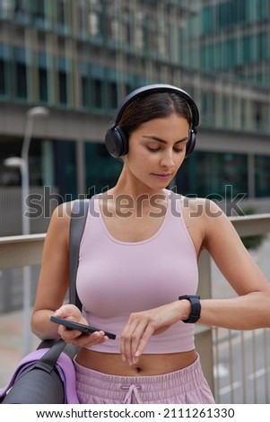 Vertical shot of active sporty woman dressed in tracksuit focused at smartwatch uses sport app on cellular listens music via headphones carries rolled mat stands outdoors against blurred background