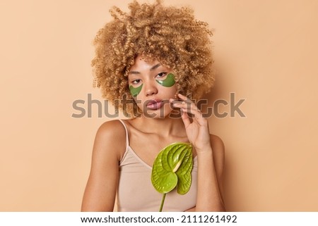 Portrait of tender charming touches face has natural beauty applies green collagen patches under eyes to remove puffiness wears casual t shirt looks directly at camera. Skin treatment concept