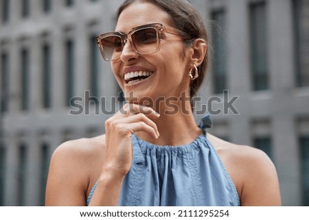 Happy young brunette European woman holds chin looks away smiles broadly being in good mood wears trendy sunglasses and fashionable clothes poses against blurred background spends free time in city