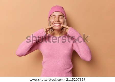 Happy millennial girl points at toothy smile keeps eyes closed shows perfect white teeth after whitening wears pink hat and jumper isolated over brown background feels positive and carefree.