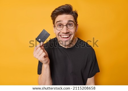 Financial services concept. Positive brunet adult man holds credit card uses electronic money happy to get money on account wears round spectcles and black t shirt isoated over yellow background.