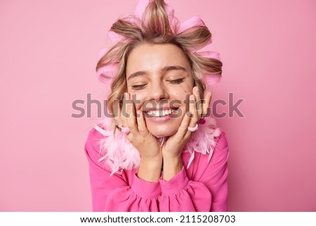 Portrait of happy young woman keeps hands on cheeks smiles broadly keeps eyes closed from satisfaction wears hair rollers for making hairstyle prepares for party isolated over pink background