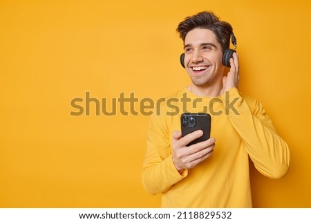 Sincere handsome man smiles gladfully wears headphones on ears listens favorite music uses cellphone dressed in casual jumper looks away being in good mood isolated over yellow studio background