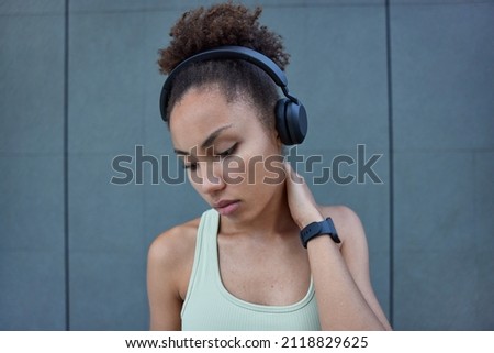 Tired sporty sweaty woman keeps hand on neck feels exhausted after sport training dressed in t shirt looks down listens music via headphones poses against grey wall wears wristwatch. Physical activity