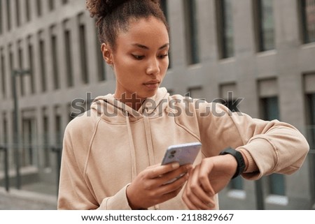 Cropped shot of sporty young woman downloads sport application for tracking fitness results holds cellphone and looks at smartwatch wears casual hoodie poses against blurred background outdoors