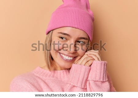 Headshot of beautiful dreamy European woman keeps hands near face smiles happily concentrated away wears pink hat and comfortable jumper has pleasant thoughts isolated over brown background.