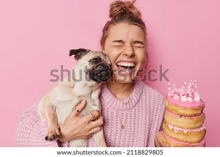 Joyful woman has fun with pug dog has upbeat mood holds festive delicious donuts celebrate pet birthday wears knitted sweater isolated over pink background. Special occasion and celebration.