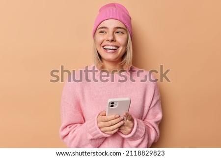 Cheerful young woman uses mobile phone sends text messages looks away has broad smile orders something in online store wears pink jumper and hat isolated over brown background. Technology concept