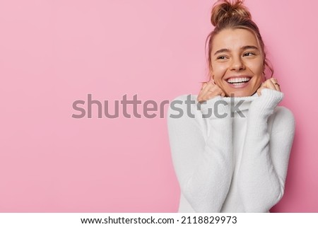 Joyful pretty young European woman with combed hair keeps hands on collar of white soft jumper smiles roadly being in good mood admires something isolated over pink background blank copy space