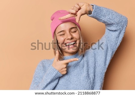 Happy young woman smiles broadly tries to find perfect angle makes frame gesture over face with fingers makes memorable photo wears pink hat casual blue jumper poses against brown background