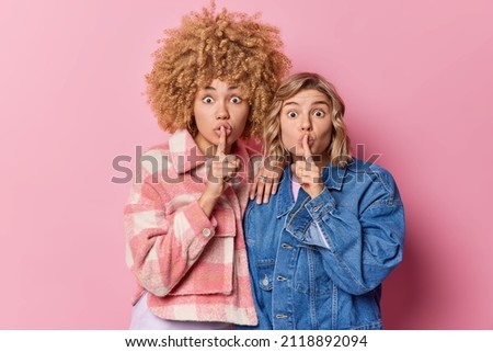 Its secret. Surprised mysterious young women make silence sign keep index fingers over lips show shh gesture look shocked at camera dressed in fashionable jackets isolated over pink background