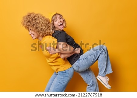 Positive friends give piggyback ride for each other foolish around dressed in casual clothes laugh joyfully isolated over yellow background feel lively and energetic. Two wmen have fun indoor