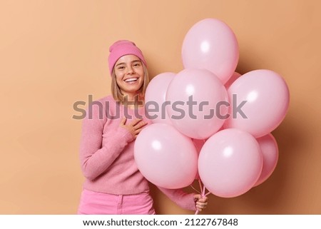 Happy millennial girl keeps hand on chest expresses gratitude celebrates anniversary poses with bunch of pink inflated balloons dressed casually isolated over brown background. Party time concept