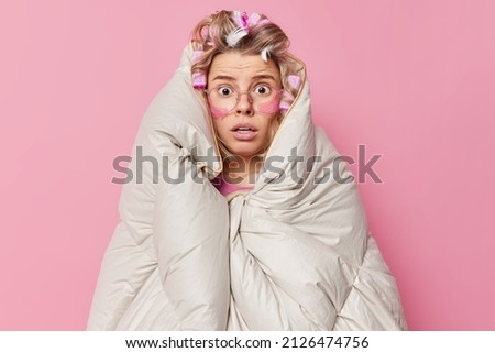 Stupefied scared woman wrapped in warm blanket stares at camera wears hair rollers big transparent glasses for good vision undergoes beauty treatments appliess patches under eyes poses indoor