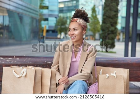 Positive curly haired woman shopper rests after making purchases in mall dressed in fashionable clothes surrounded by shopping bags looks gladfully away poses against blurred urban background.