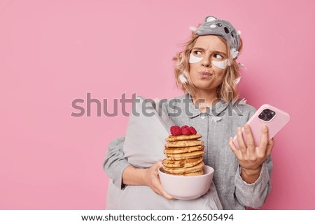 Unhappy annoyed woman looks angrily away makes plannings in mind uses mobile phone for surfing internet holds bowl of pancakes with raspberries applies beauty patches holds pillow stands indoor
