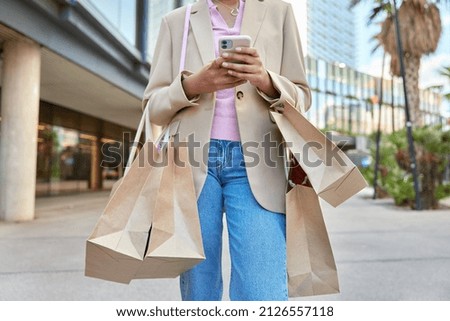 Cropped shot of unrecognizable woman walks outdoors with shopping bags returns from mall wears fashionable jacket and jeans poses in city centre holds smartphone messages after making purchases