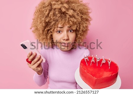 Indignant puzzled curly haired woman holds mobile phone and heart shaped cake prepares for celebration of special occasion waits forr guests poses against pink background. Happy birthday concept Stock fotó © 