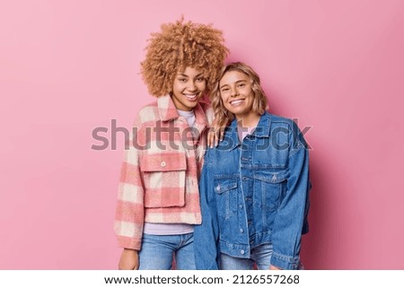 Satisfied young women smile pleasantly stand next to each other dressed in fashionable jackets being in good mood isolated over pink background. Two joyful female friends meet together pose for photo
