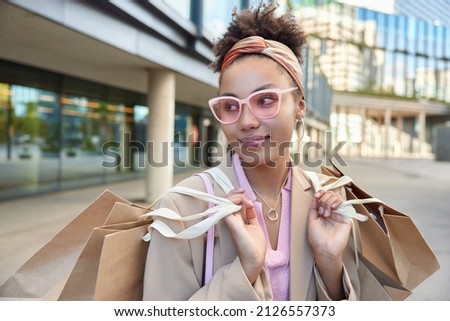 Horizontal shot of fashionable female shopper wears trendy pink sunglasses headband carries paper bags after making shopping walks outside during sunny weather. Consumerism sale purchases concept