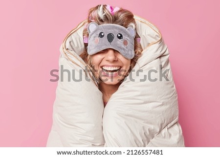 Unrecognizable cheerful woman wears sleepmask on eyes smiles broadly wrapped in duvet expresses positive emotions being in good mood isolated over pink background. People rest and bedtime concept