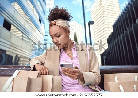 Outdoor shot of stylish woman looks attentively in shopping bags looks through her purchases uses smartphone poses on bench wears fashionable clothes against urban background. Consumerism concept