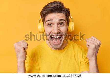 Energetic positive adult man clenches fists feels like winner or champion wears stereo headphones on ears being full of happiness enjoys listening favorite music isolated over yellow background.