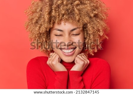 Close up portrait of cheerful curly haired young woman keeps hands on collat of turtleneck closes eyes from satisfaction expresses positive emotions smiles toothily isolated over red background