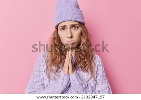 Sad young woman with imploring look keeps palms pressed together begs for something needs your help and support wears purple loose jumper and hat isolated over pink background. I am so sorry Imagine de stoc © 