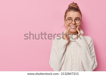 Horizontal shot of pretty cheerful woman with bun hairstylepoints index fingers at smile shows white teeth concentrated away wears spectacles and white knitted sweater isolated on pink background