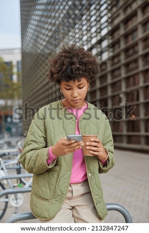 Outdoor shot of curly haired young woman drinks aromatic coffee and surfs internet on mobile pone wears jacket watches video online spends free time in city poses against blurred background.
