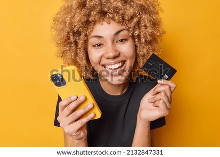 Happy curly female shopper enjoys easy paying for goods online smiles broadly shows white teeth uses mobile phone and credit card dressed in casual black t shirt isolated over yellow background