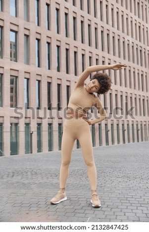 Full length shot of active slim curly haired woman dressed in tracksuit leans away exercises outdoors has self confident expression poses near modern building has regular workout during morning.
