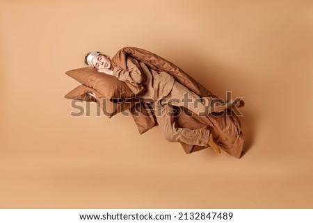 Relaxed peaceful young European woman leans on pillow lies under blanket sleeps and sees sweet dreams wears sleepmask and pajama flies in air isolated over brown background. Good night concept