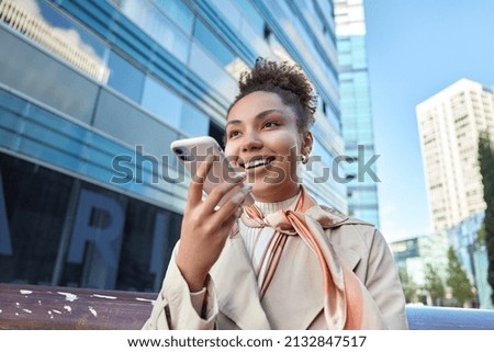 Glad beautiful young woman dressed in coat and kerchief around neck records voice call holds smartphone near mouth poses against modern city background. Modern technologies and lifestyle concept