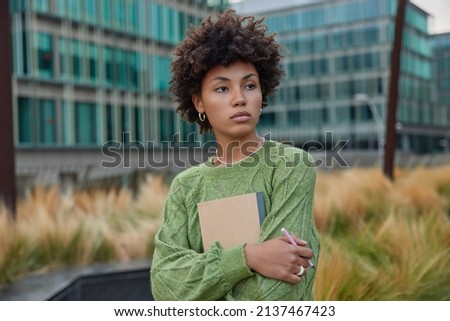 Beautiful curly woman holds notepad and pen concentrated into distance focused into distance thinks about creative ideas wears casual green jumper poses outdoors against blurred urban background