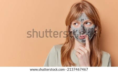 Serious pensive teenage girl with ginger hair looks away thinks how to improve skin condition applies clay mask on face isolated over beige background copy space for your promotion. Beauty concept