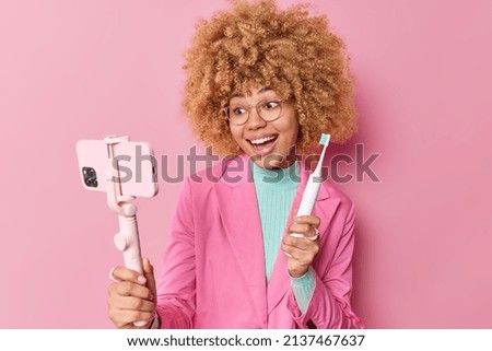 Oral care and hygiene concept. Cheerful woman records video for personal blog advertises electric toothbrush tells how to care about teeth holds mobile phone on stick isolated over pink background Stock foto © 