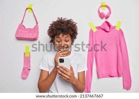 Positive curly haired young woman uses smartphone surfs internet reads receieved message dressed in casual basic t shirt poses against white background with plastered pink items to wear around.