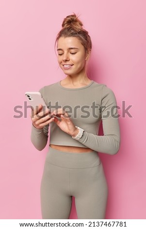 Vertical shot of slim sporty woman dressed in tracksuit surfs social networks via smartphone rests after training in gym poses against pink background uses fitness app chooses music for workout